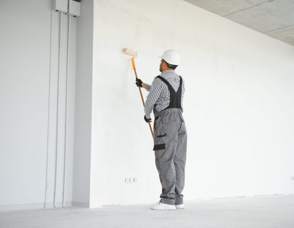 An Indian apartment repair worker paints a white wall with a roller.
