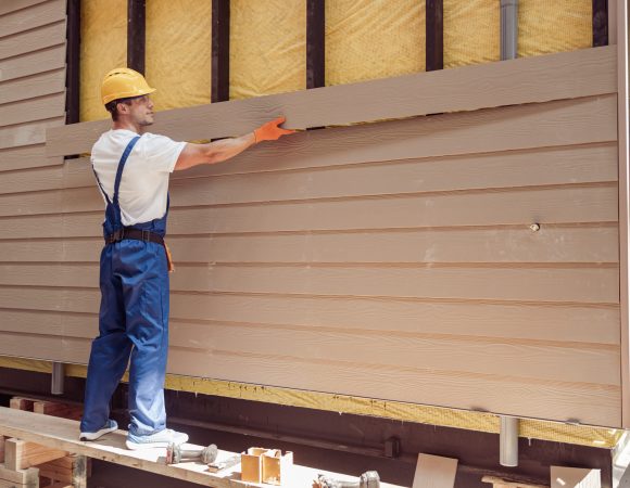 Smiling young man construction worker wearing safety helmet and work overalls while installing exterior wood siding